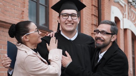 Graduation from university: student hugs his parents, they are happy and proud of their son. Wearing black costume - mantle and hat. Getting high education, celebrating graduation.