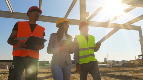 Group of builders and architect walking on construction site. Teamwork - female architect, construction engineer and manager with tablet and clipboard talking about the project with bright sun rays