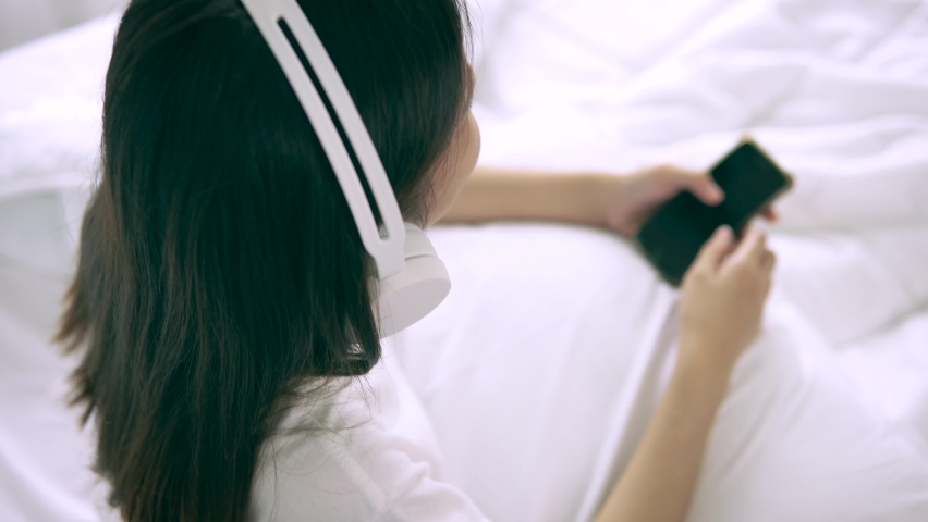 Happiness young woman with headphones relaxing at the bed using mobile phone to choose music and enjoy listening music at home | Shutterstock HD Video #1060343750