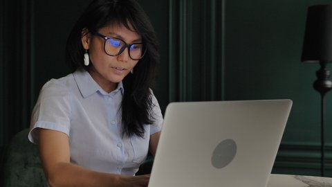 Experienced businesswoman in white blouse and glasses opens grey laptop and types sitting at table against green wall slow motion