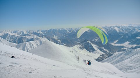 Beautiful epic landscape of high altitude mountains, bright coloured paraglider parachute flies down. Amazing scene of extreme sports for winter activity at ski resort