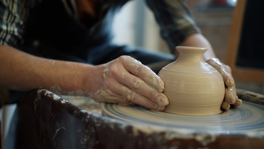 Close-up of potter's hands covered with clay making beautiful vase on throwing wheel in pottery workshop. Creativity and traditional crafts concept. Royalty-Free Stock Footage #1060353470