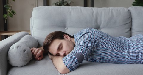 Exhausted unmotivated young european man falling asleep on couch, feeling no energy after hard working day. Overwhelmed 30s businessman flopping down on sofa, needs rest at home, burnout concept.