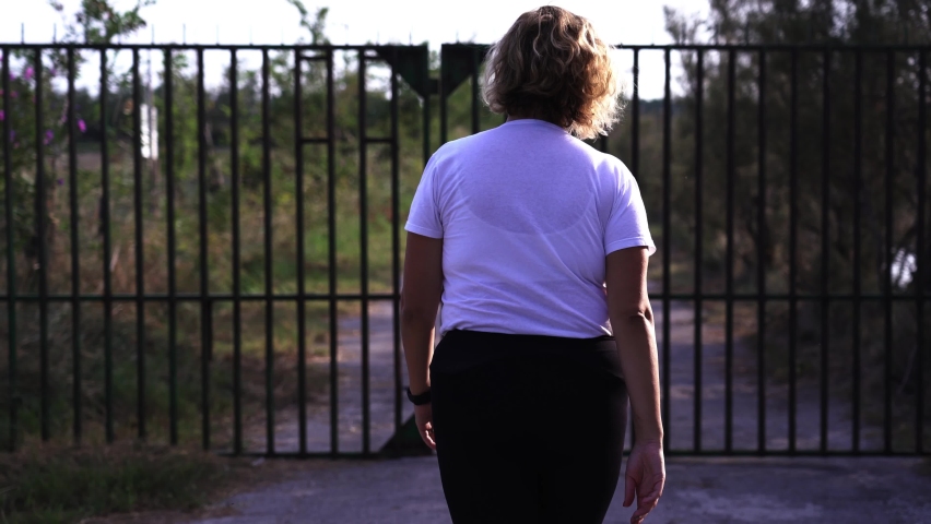 Blonde with short curly hair walks along road to metal fence