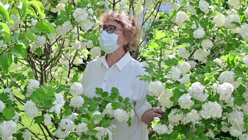An elderly woman in a protective mask sniffs a blossoming apple tree