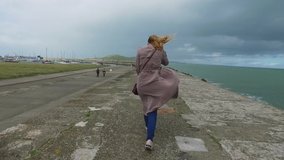  View from the back of girl walking on pier in bad weather. Strong wind blows down young woman. The fabric of coat and scarf flutters. Storm at sea in Ireland near Dublin. High quality FullHD footage