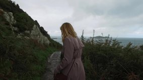 The girl walks down the hill. View of a young woman from the back. The fabric of the long coat flutters. Tracking in Ireland near Dublin city. High quality FullHD footage video.