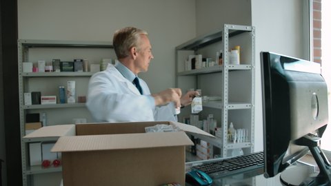 Pharmacist unpacking delivery from stock indoor of drug store interior. Caucasian mature man fills the shelves with medicine pills, ampoules of saline etc. Middle view, daylight.