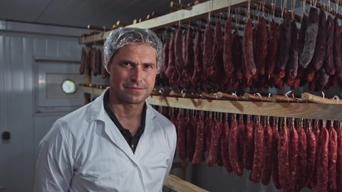 Portrait of meat expert worker handsome man in smokehouse. Racks of salami sausage sticks hanging on background. Meat factory.