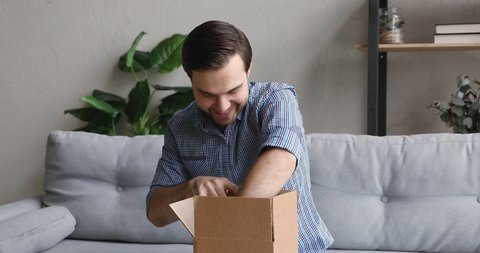 Curious young handsome smiling man client sitting on sofa at home, unpacking small carton box with wished item, satisfied with fast delivery service, having positive internet shopping experience.