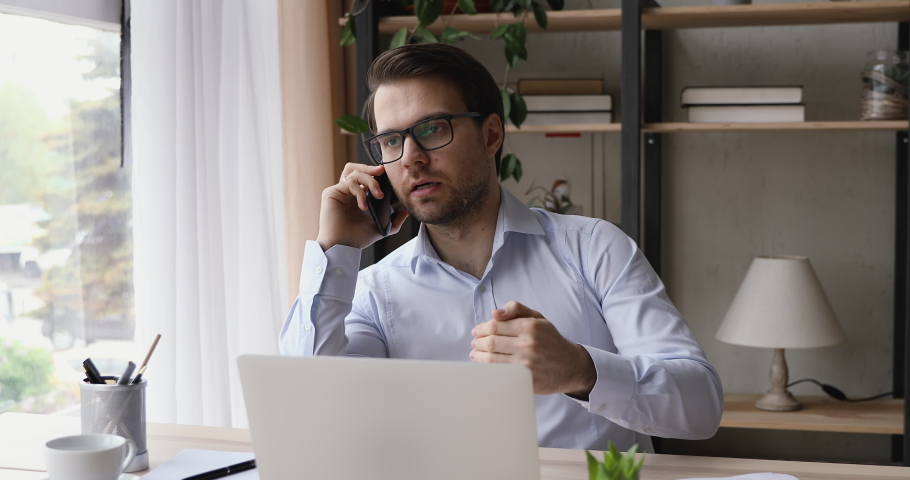 Happy young businessman in eyeglasses sitting at table with computer, holding mobile phone call with clients or partners, discussing working issues, negotiating project, enjoying conversation. Royalty-Free Stock Footage #1060360703