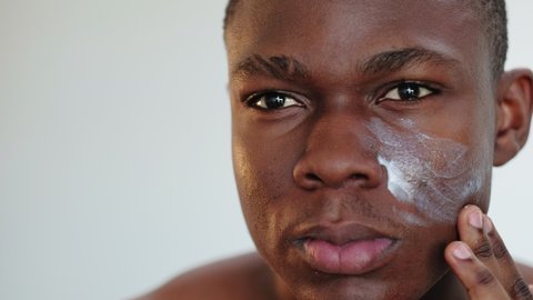 Man skincare. Acne treatment. Portrait of satisfied african guy applying cream on face smiling isolated on light copy space background. Dermatology hygiene.