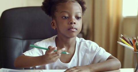 Close up portrait of concentrated little African girl learning drawing painting picture with colored pencils. American female pupil kid looking at laptop screen, child home online remote education