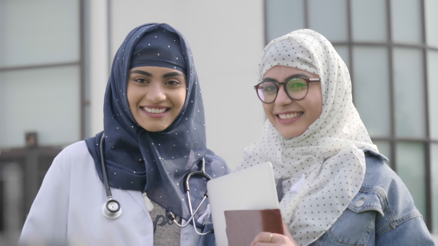 Two attractive Muslim females doctors or physicians wearing headscarves and aprons are smiling. Two happy Islamic undergraduate girl medical students or interns are standing outside a hospital Royalty-Free Stock Footage #1060364900