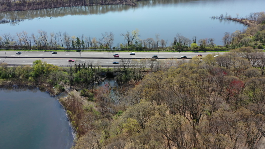 An aerial view of some reflective lakes during a sunny day. The drone truck left along side the parkway & tilts down. It flies over one of the lakes as it observes the traffic on the road.