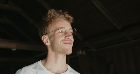 Unshaven blond man wearing glasses smiling to himself inside a wooden building before running his hand through his tousled hair with a beaming smile of satisfaction