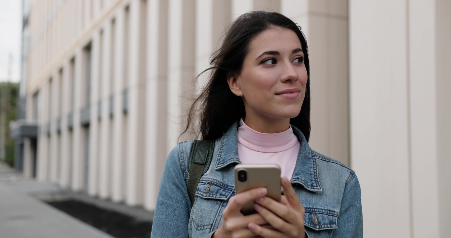 Close up portrait of cheerful young Caucasian female standing on street and tapping on smartphone. Beautiful joyful woman holding mobile phone in hands outdoors and smiling to camera. Urban concept | Shutterstock HD Video #1060379978