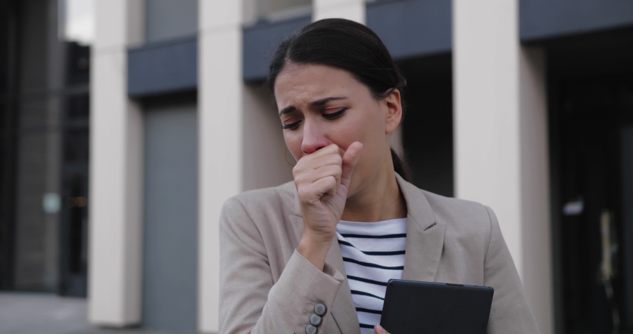 Close up portrait of beautiful Caucasian woman walking in city on street with tablet in hand and covering face with hand while coughing. Sick young female sneezing and coughing. Job concept | Shutterstock HD Video #1060380011