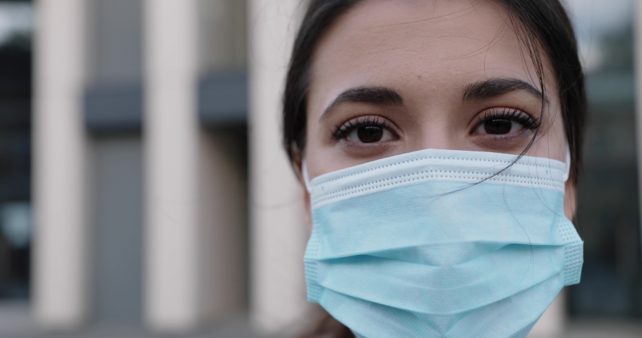 Close up of cheerful Caucasian young dark-haired female standing outdoor in medical mask on face and looking at camera. Happy pretty woman alone on street in town in quarantine. Health concept Royalty-Free Stock Footage #1060380065