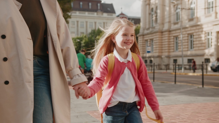 Happy excited child girl with schoolbag holding mom in hands going to school together in sunny morning. First day of school. Education, kids and emotions Royalty-Free Stock Footage #1060384235