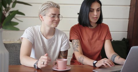 Romantic lesbians laugh and talk looking into white laptop display on brown wooden table with coffee cups in kitchen slow motion