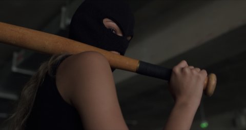 Naughty girl in a balaclava holds a baseball bat in her hands and poses on the territory of a car parking. Cheeky sexy woman in mask shows disrespect and aggression. Back view