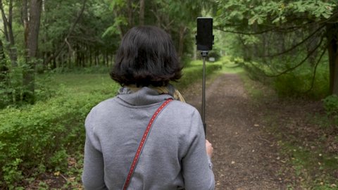 A woman with black hair in a gray sweater walks through the forest and shoots a video on her smartphone. She is a video blogger and tells her followers about different beautiful places.