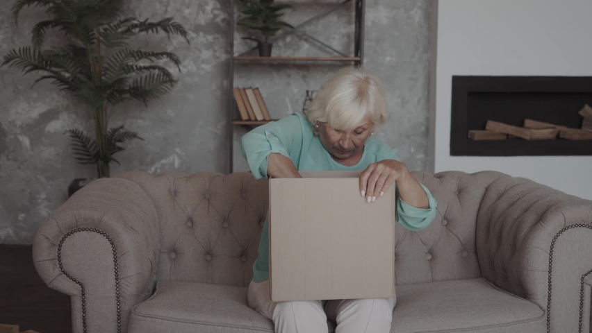 Elderly lady upset with an online purchase. A gray-haired woman pulling goods from cardboard shipping box and looks unhappy and disappointed Royalty-Free Stock Footage #1060394465