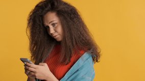 Pretty positive brunette girl looking in camera and smiling chatting with boyfriend on smartphone isolated on yellow background