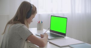 e-learning for schoolers by internet, girl is drawing in copybook listening teacher by videochat, green screen on laptop