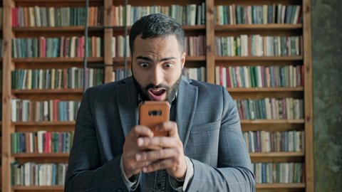 Young Man In The Library With Mobile Phone In His Hands. Guy With Beard And Black Hair. He Is Emotionally Playing Game On The Phone. Cute Young Man From The Middle East. Stylish Guy With Beard In