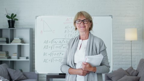 Female Math Teacher Standing On Blackboard Background With Math Formulas. Pretty Middle Aged Woman With Glasses. She Conducts Online Training Or Records Video Lesson. She Smiles. Woman Standing With