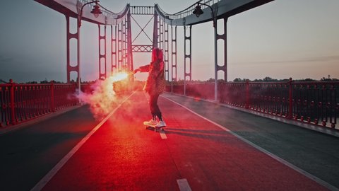 Young hipster female with pink hair, in informal outfit is skateboarding on deserted bridge holding burning red signal flare and waving it. Early morning, tracking shot. Back view, slow motion