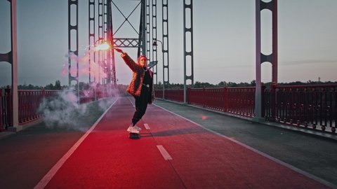 Young hipster lady with pink hair, in informal outfit is riding skateboard on deserted bridge while holding glowing red signal flare and waving it. Early morning, tracking shot. Slow motion