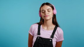 Pretty young girl with long hair listening to music, smiling, dancing head in pink headphones on studio against blue background. Music, dance, radio concept, slow motion.