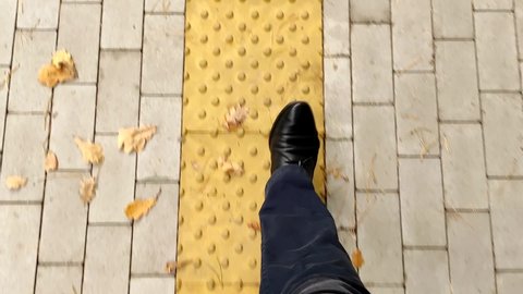 View from the top of the legs of a man walking on a tactile warning tile for blind people. A handicapped person walks along the tactile tile sidewalk. Tactile pedestrian sidewalk for blind people.