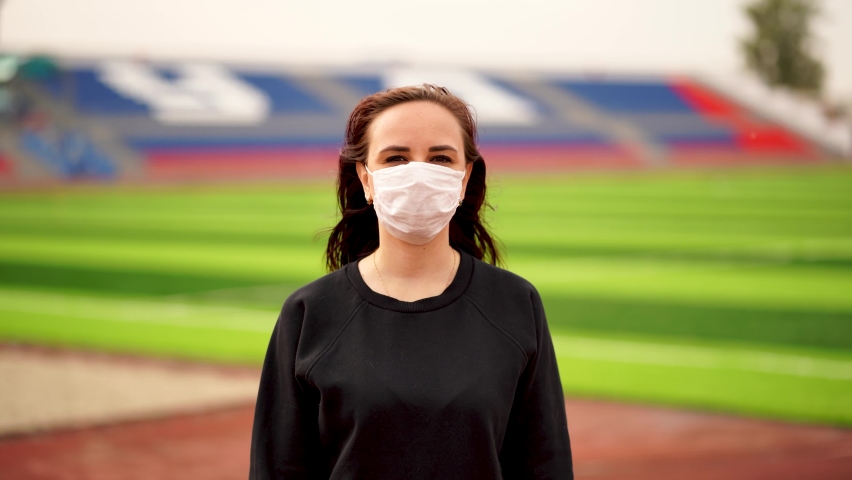 Young woman in medical mask doing warm-up at stadium. Adult female in black casual clothes doing sports in fresh air and protecting yourself from diseases in public place.