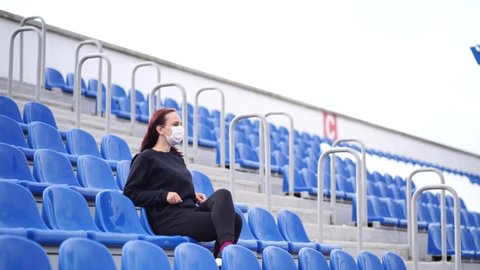 Young woman in medical mask looks away, sitting on stadium bleachers alone. Adult female in black casual clothes protecting yourself from diseases. Concept of threat of coronavirus epidemic infection.