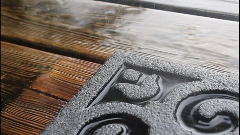 Raindrops falling and splashing on a wooden deck and doormat at an entryway to a home.