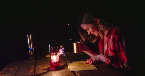 Young woman in red classic shirt sitting at wooden table by campfire looking on the map and drinking hot tea enjoying outdoor adventure. Red lantern lightning the night scene. Woman on camping