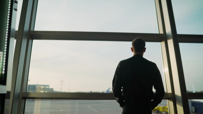 A respectable young man stands at the airport and looks at the plane. Young guy at the airport. The guy is standing at the large window at the airport. He looks outside and watches the plane Royalty-Free Stock Footage #1060415149