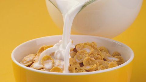 Milk pouring into bowl of corn flakes with splashing in slow motion, healthy cereal breakfast on yellow background
