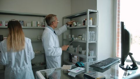 Two colleagues pharmacists checking up medicine stuff in drug store. Adult senior male in white robe and young caucasian woman rowing correctly pills, ampoules and cream.