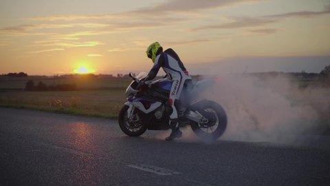 Lubliniec , Lubliniec / Poland - 10 05 2020: Medium wide slow motion shot of a motorbiker wearing full body suit and helmet performing a burnout with a white superbike during sunset