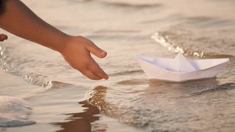 Hand of a little girl lowers a paper boat into the river. Children's paper boat on waves of the river. Close-up of a little girl's hand with a paper boat. Child playing on the river beach at sunset