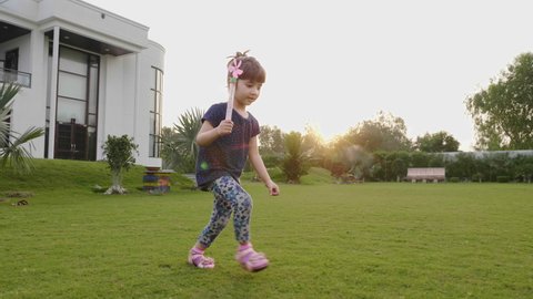 A happy and smiling young adorable or cute little girl child is running with a pinwheel on the grass or lawn in the garden or park against the sun. 
