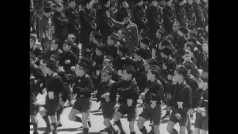 1940s: Children march in uniforms. Two girls give each other salute. Children marching in uniforms. Nazi flag. Quote from Hitler. Children march in uniforms carrying Nazi flags.
