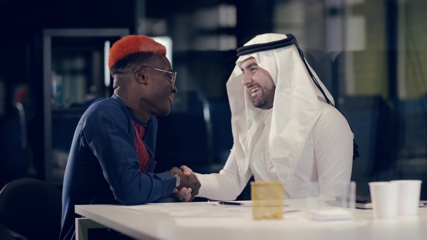 An Arab businessman in kandur and his African colleague in national dress have a business conversation while sitting at a table in a modern meeting room and come to an agreement by shaking hands. | Shutterstock HD Video #1060430815