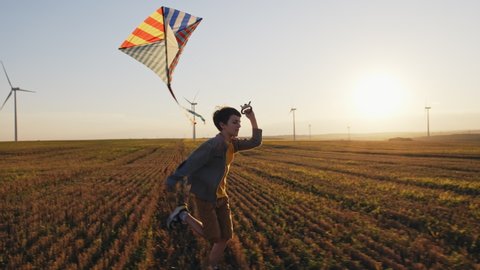Happy boy runs launches bright kite into sky mown wheat field, playing with wind in field of an orange sunset on day lens flares wind turbines in summer slow motion. School break. Lifestyle. Childhood