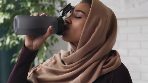 Modern young Indian woman in hijab scarf drinking water from sport bottle during workout pause at home, rehydrating, staying healthy. Active muslim lady resting after fitness training.